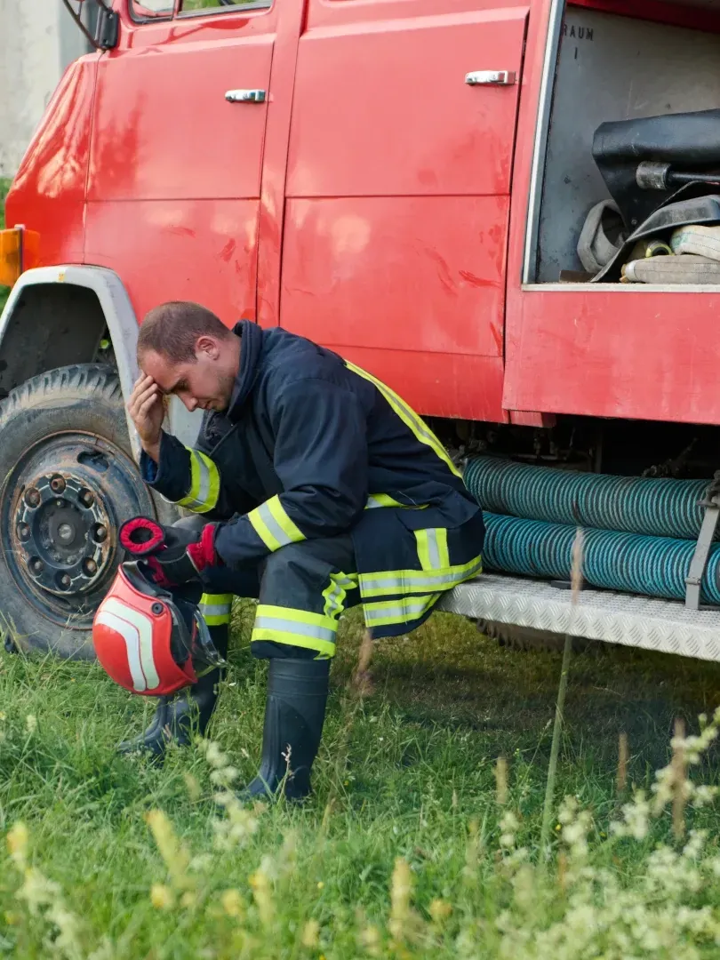 Firefighter kneeling beside a red truck, adjusting gear near the wheel on a grassy roadside.