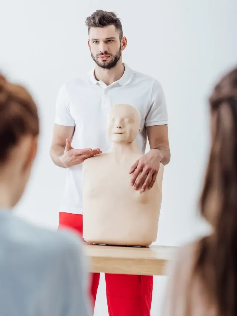 Instructor demonstrating CPR on a training mannequin in a classroom, with students watching.