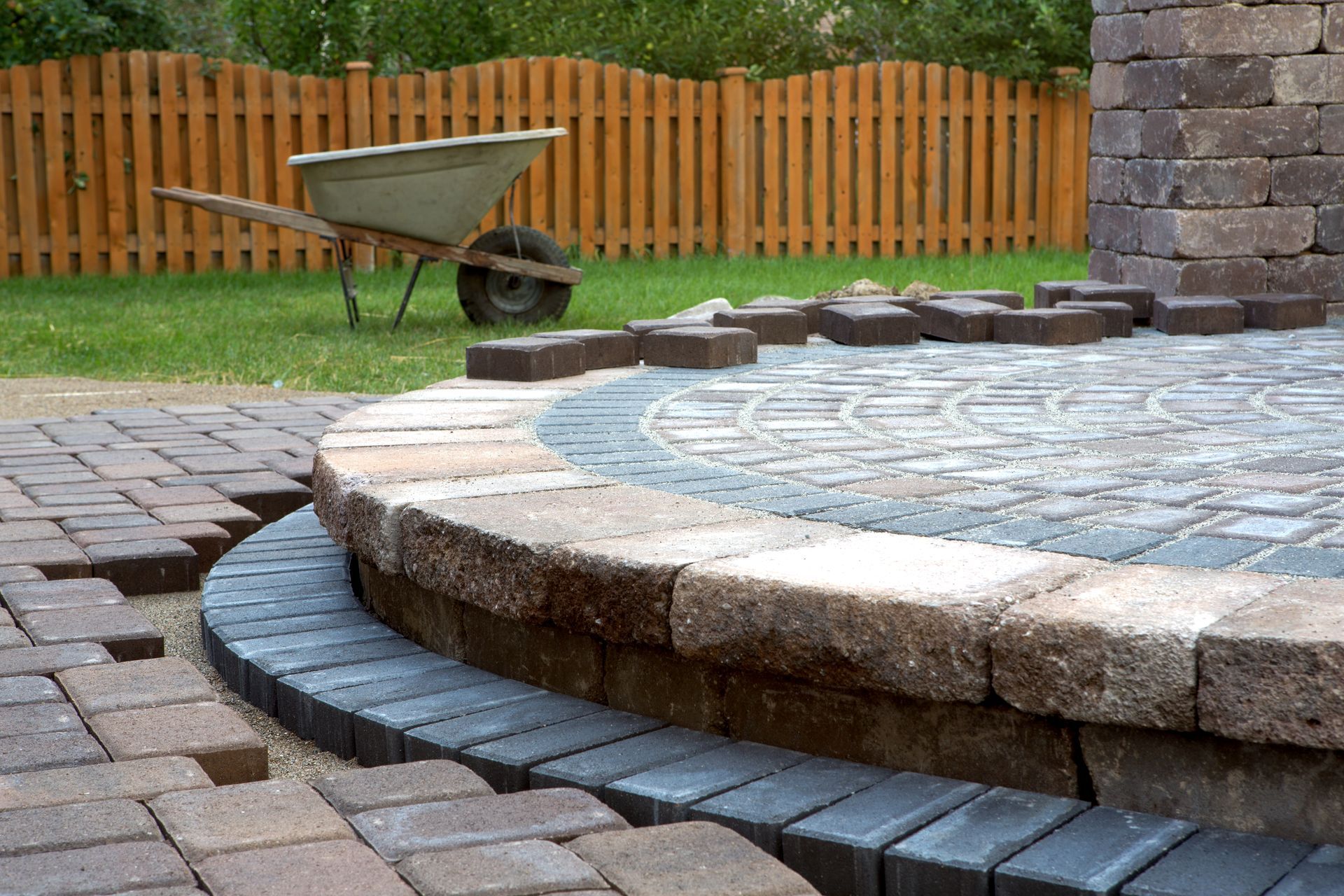 A wheelbarrow is sitting on the grass next to a brick patio being built.