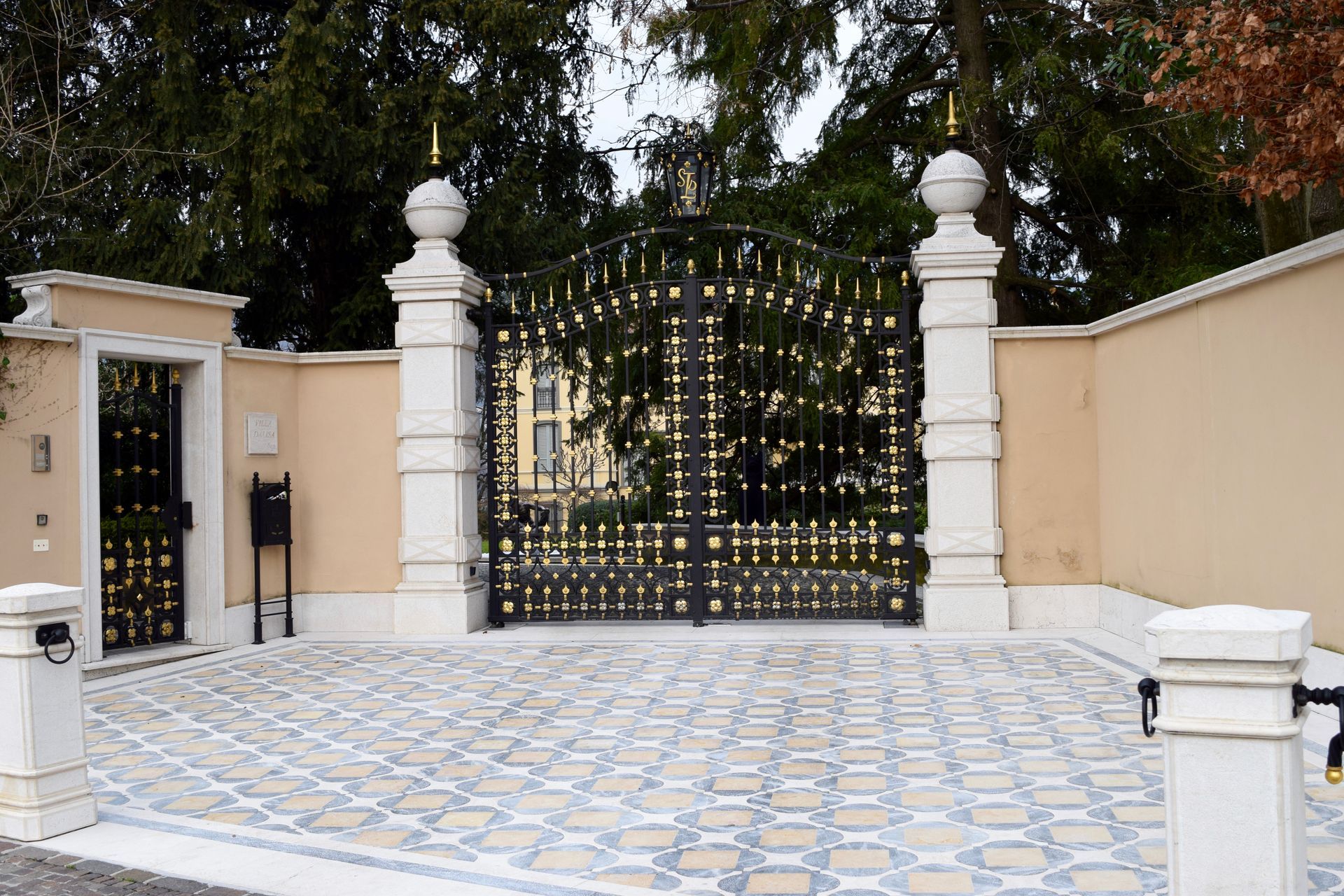 Ornate black and gold gate with pillars, leading to a driveway.