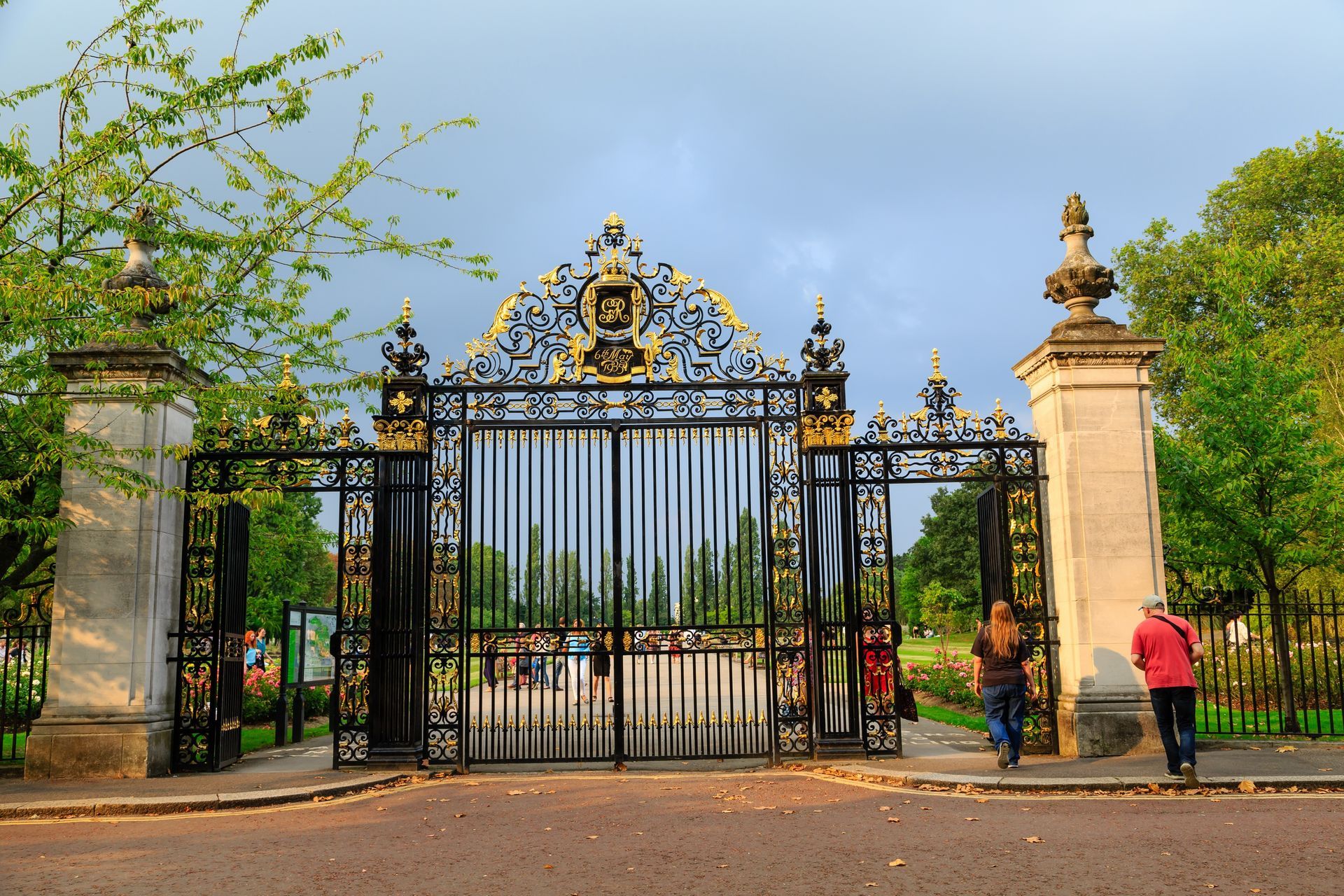 Ornate black and gold gates with stone pillars at park entrance; people walking in.