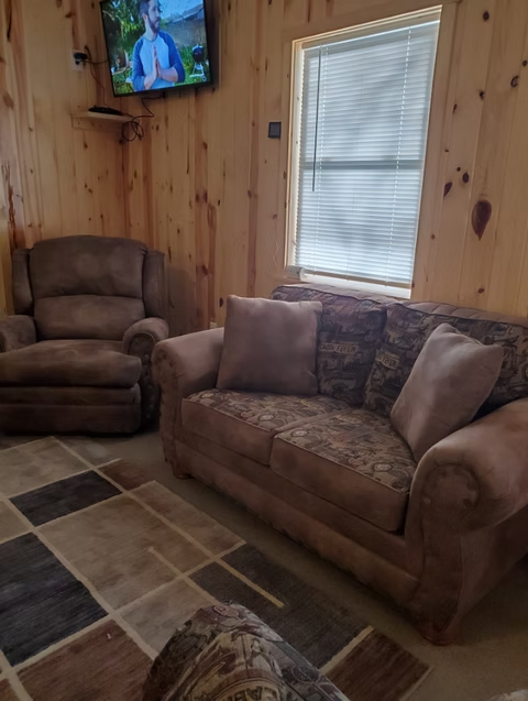 Cozy wood-paneled living room with brown sofa, recliner, window blinds, and wall-mounted TV