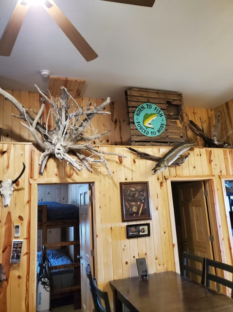 Rustic wood-paneled dining room with a mounted fish clock and ceiling fan above a table