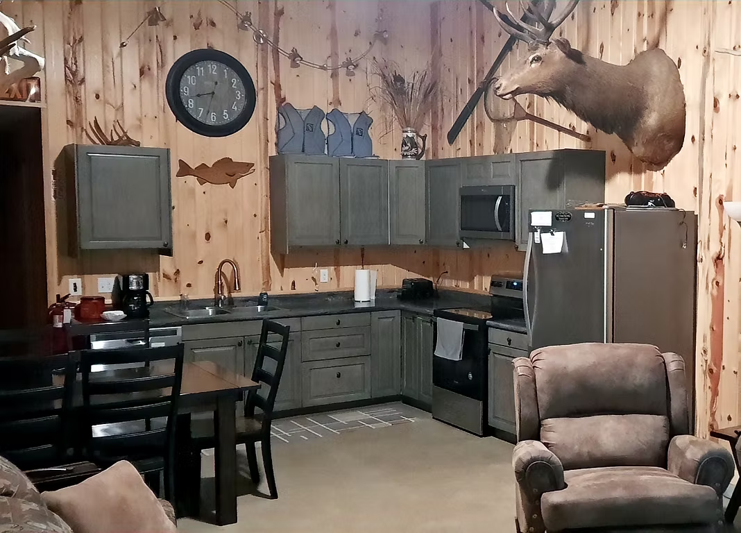Rustic kitchen with gray cabinets, wood-paneled walls, dining table, chairs, and a mounted deer head.