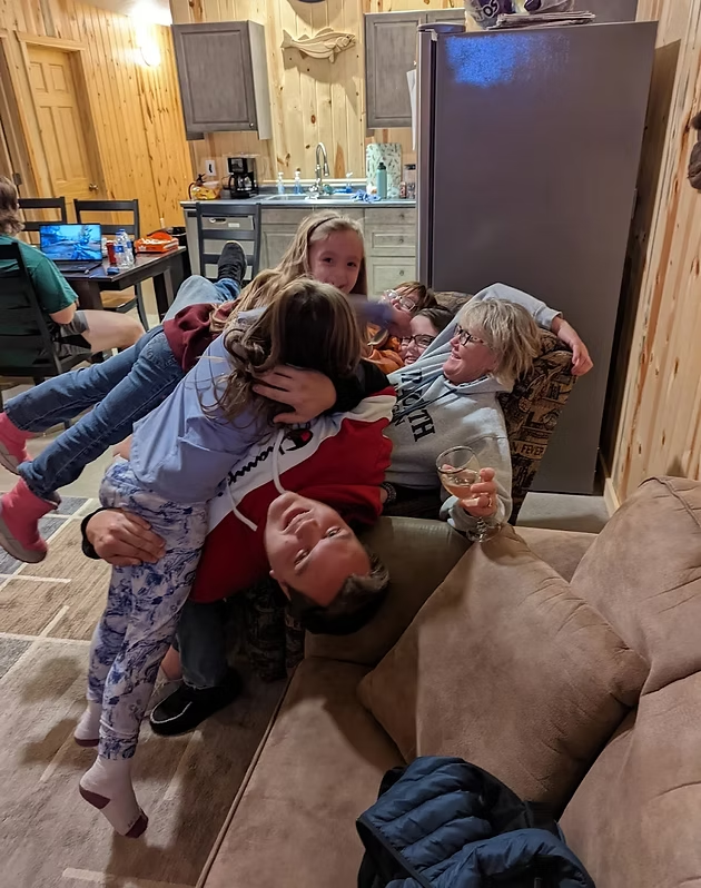 Three children in pajamas hug an adult on a couch in a cozy wood-paneled living room.