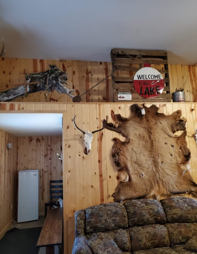Rustic wood-paneled room with mounted deer hide, antlers, camouflage decor, and a “Welcome to the Lake” sign.