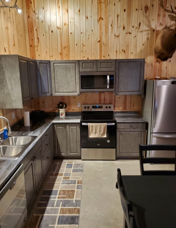 Wood-paneled kitchen with gray cabinets, stainless fridge, stove, sink, and checkered rug