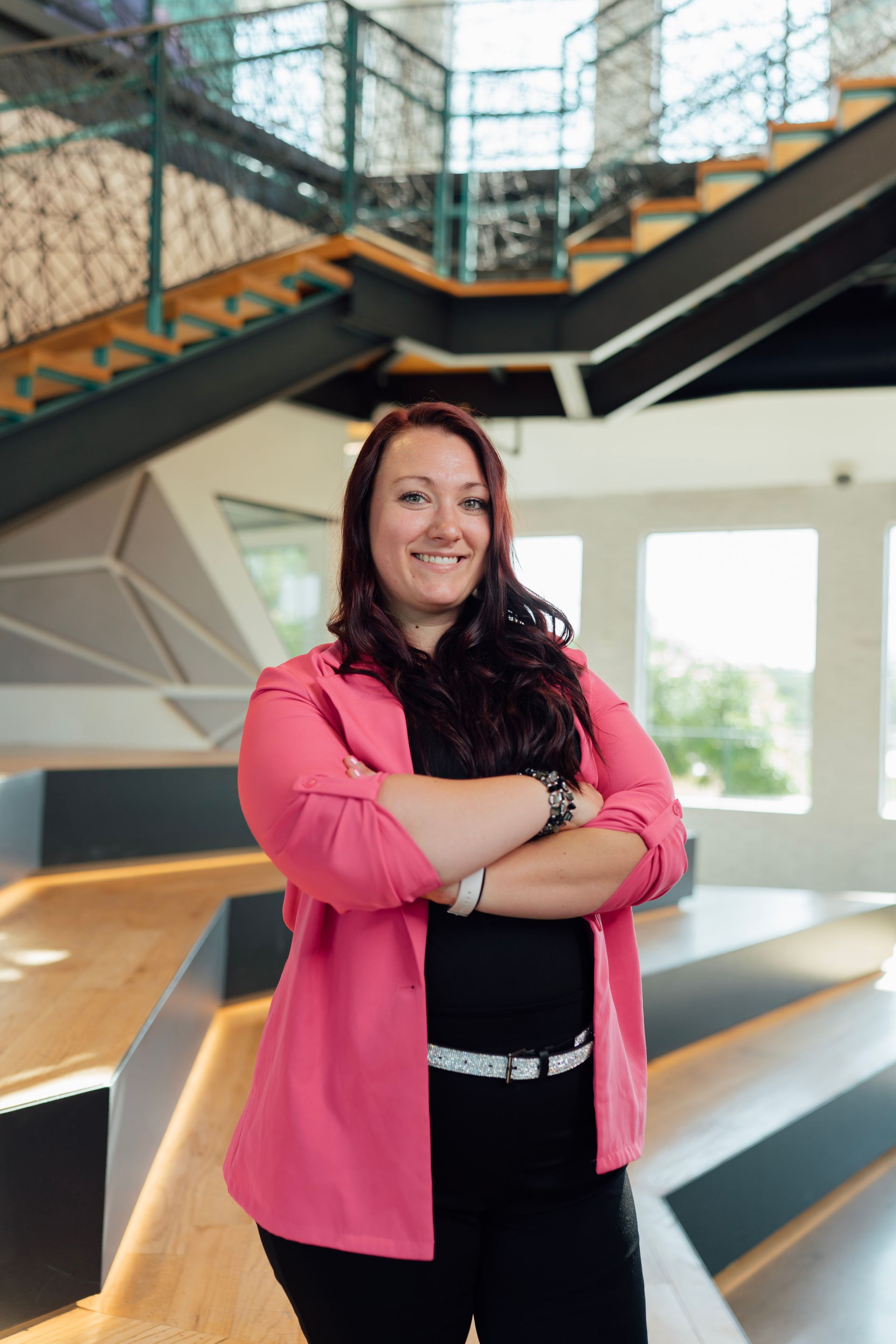Woman with crossed arms in a pink blazer, black top and pants, smiles in a modern office setting.