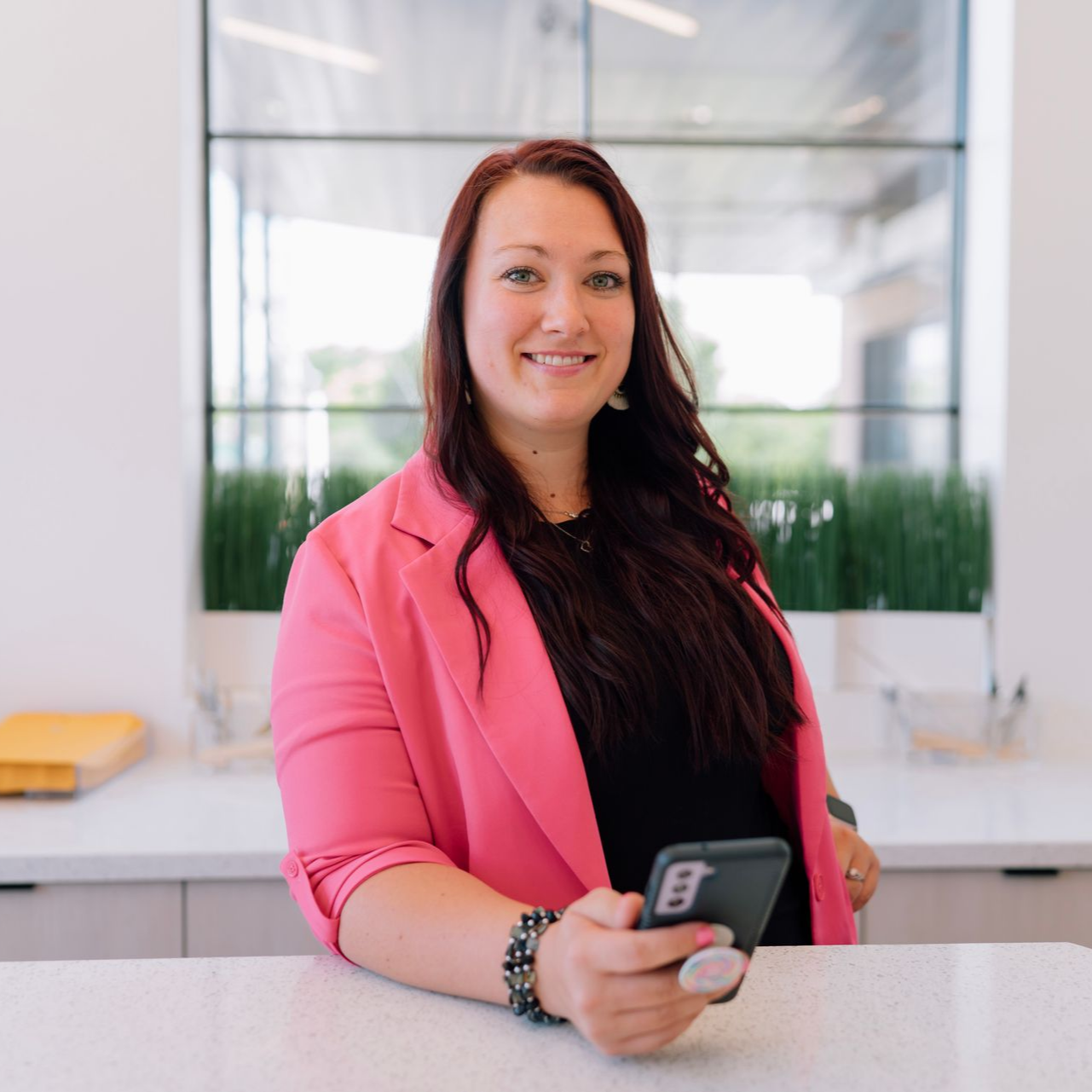 Woman in pink blazer smiles, holding a phone with a pop socket. Leaning on a counter in a modern office.