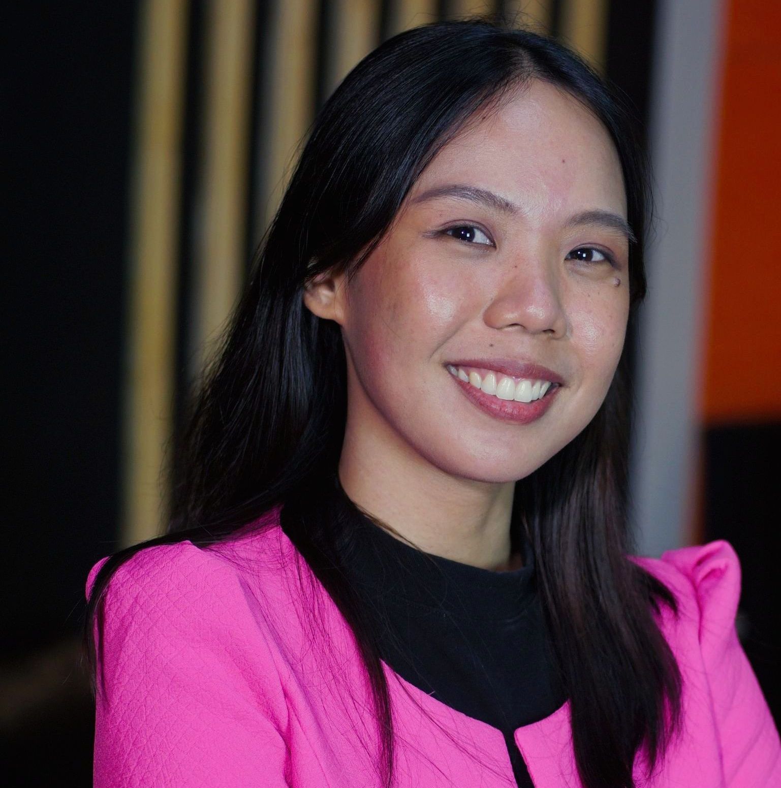 Woman with dark hair, smiling, wearing a pink blazer and black top.