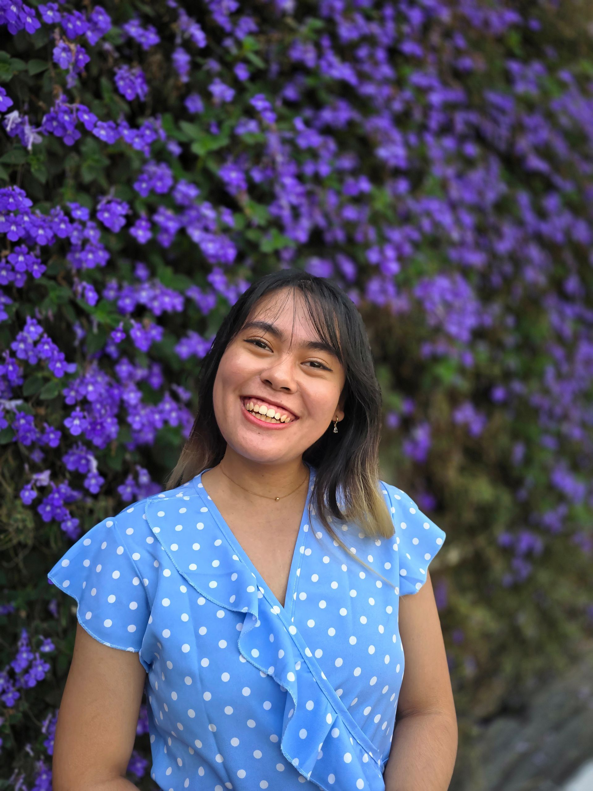 A woman in a blue polka dot shirt is smiling in front of a wall of purple flowers.