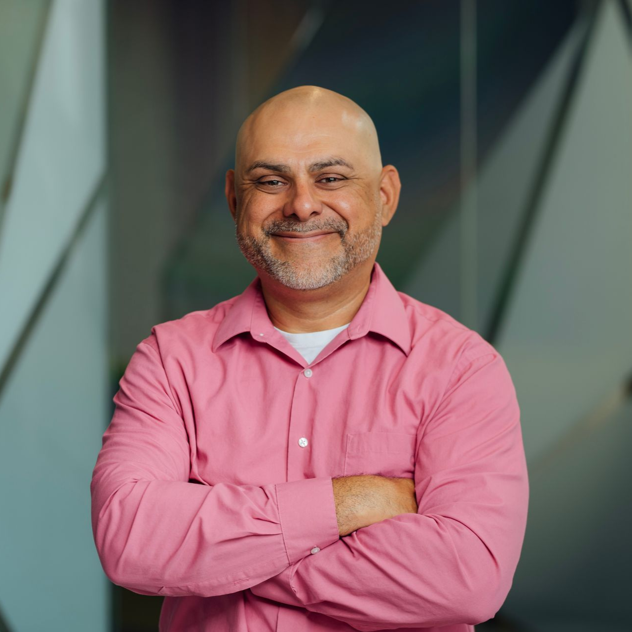 Bald man with a gray beard smiles, arms crossed, wearing a pink shirt, white undershirt, against a geometric glass background.