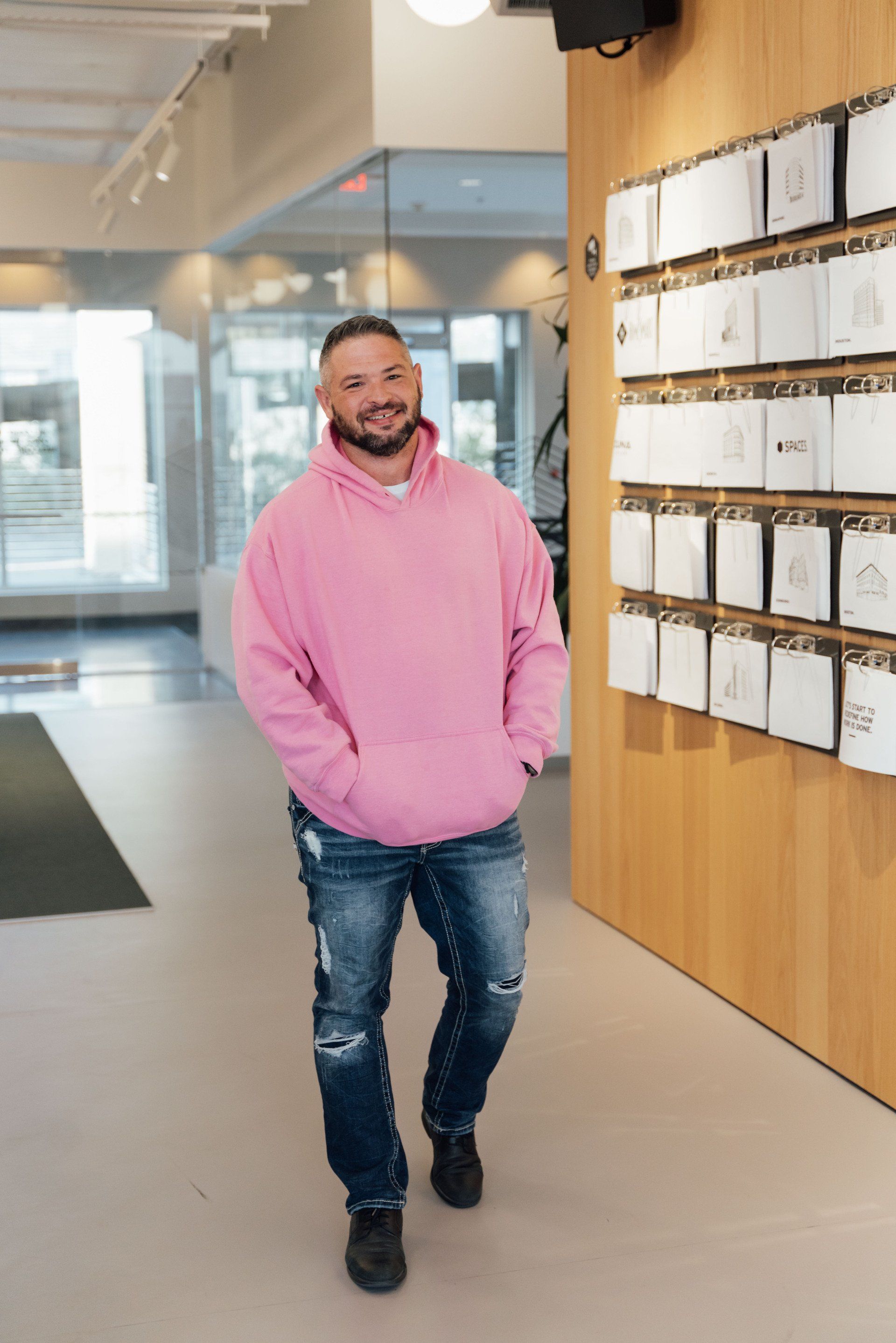 A man wearing a pink hoodie and jeans is standing in a hallway.