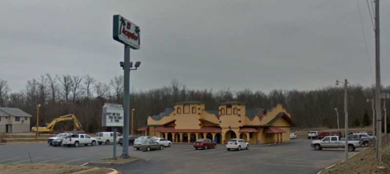A tan, multi-arched building under a gray sky, with a tall signage pole, parking lot, and construction equipment nearby.