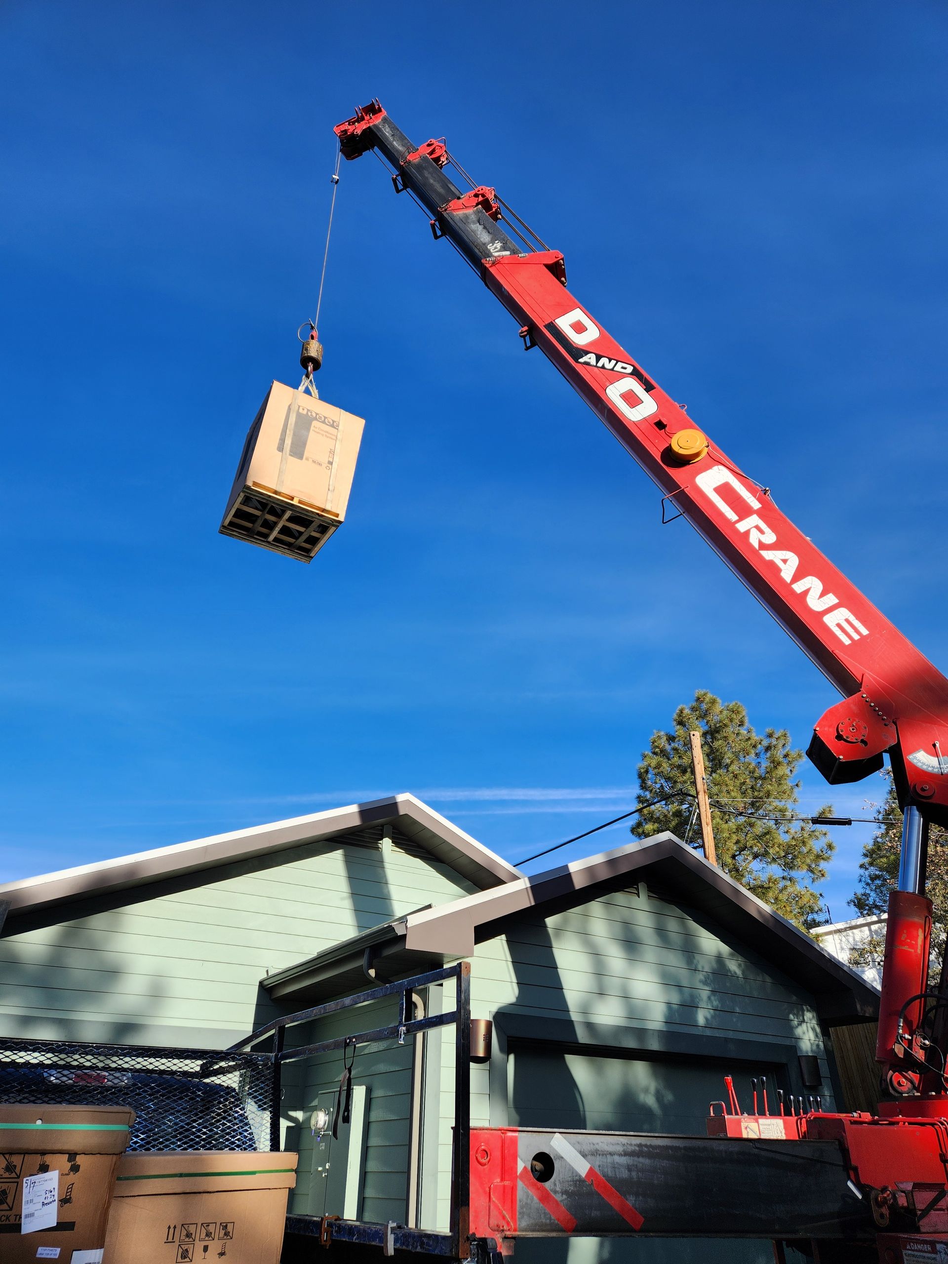 Red crane lifting a hanging crate over a house roof under a clear blue sky