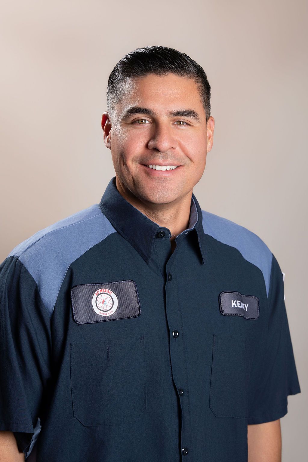 Man in dark blue work shirt with logo patches, smiling against a neutral studio background