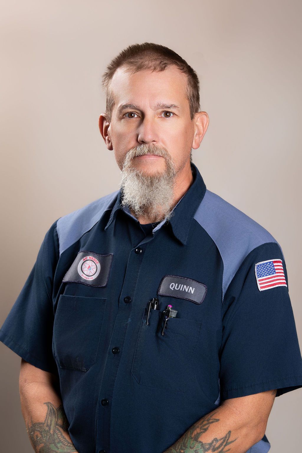 Man in a navy uniform shirt with patches stands against a plain background, looking at the camera.