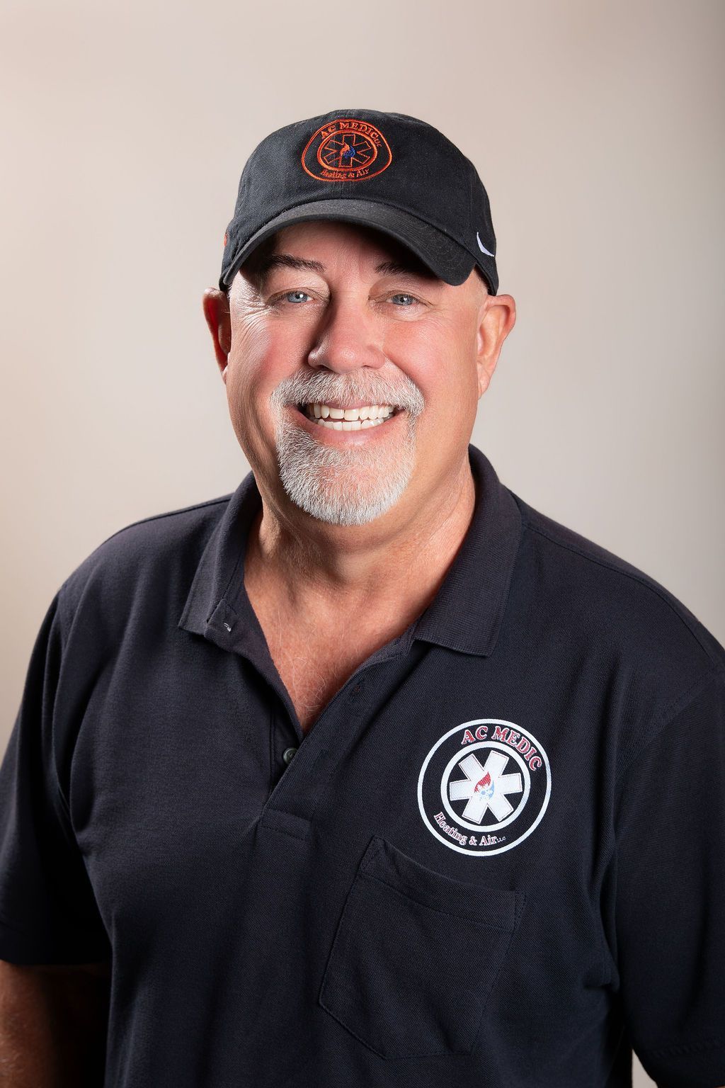 Smiling man in a black polo and cap with a circular logo, standing against a light background.