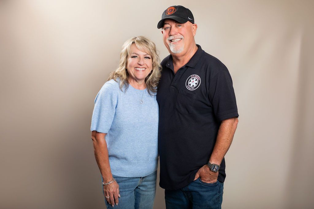 Smiling couple posing against a beige backdrop, wearing blue shirts and black cap, standing close together