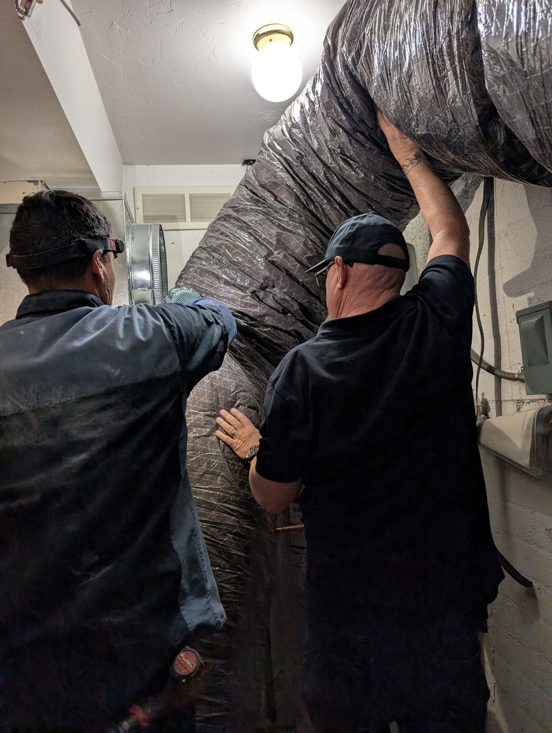 Two workers installing flexible ductwork in a basement, reaching up to secure it.
