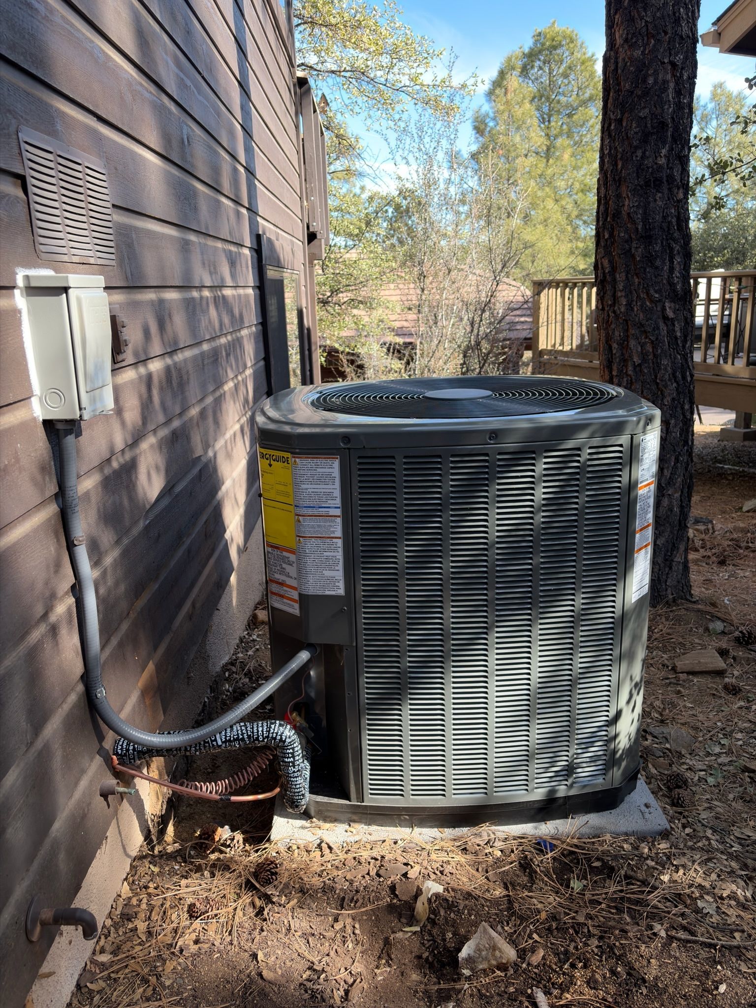 Outdoor HVAC condenser beside a house, with a window unit and tree in a wooded yard