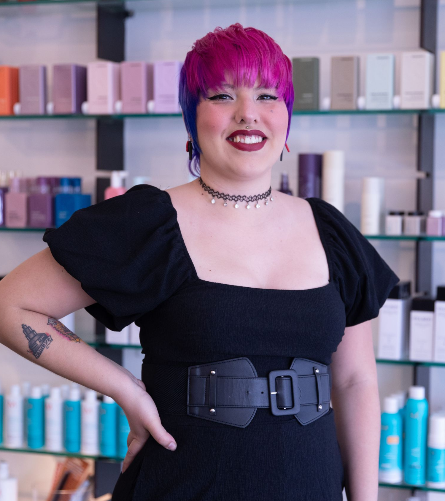 Smiling person with magenta hair in black dress and belt, standing before shelves of skincare products