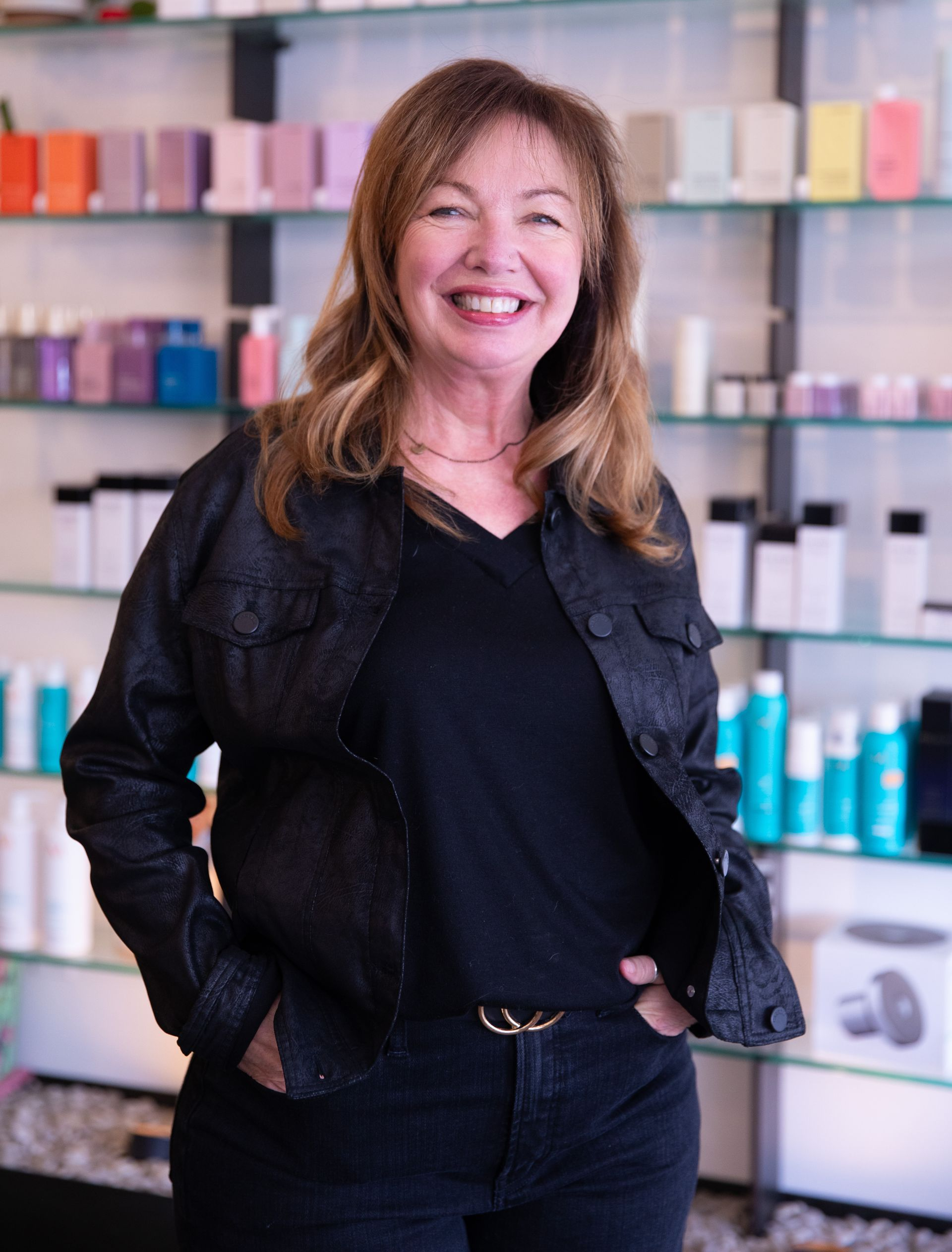 Smiling woman in black outfit standing in a salon with colorful product shelves behind her