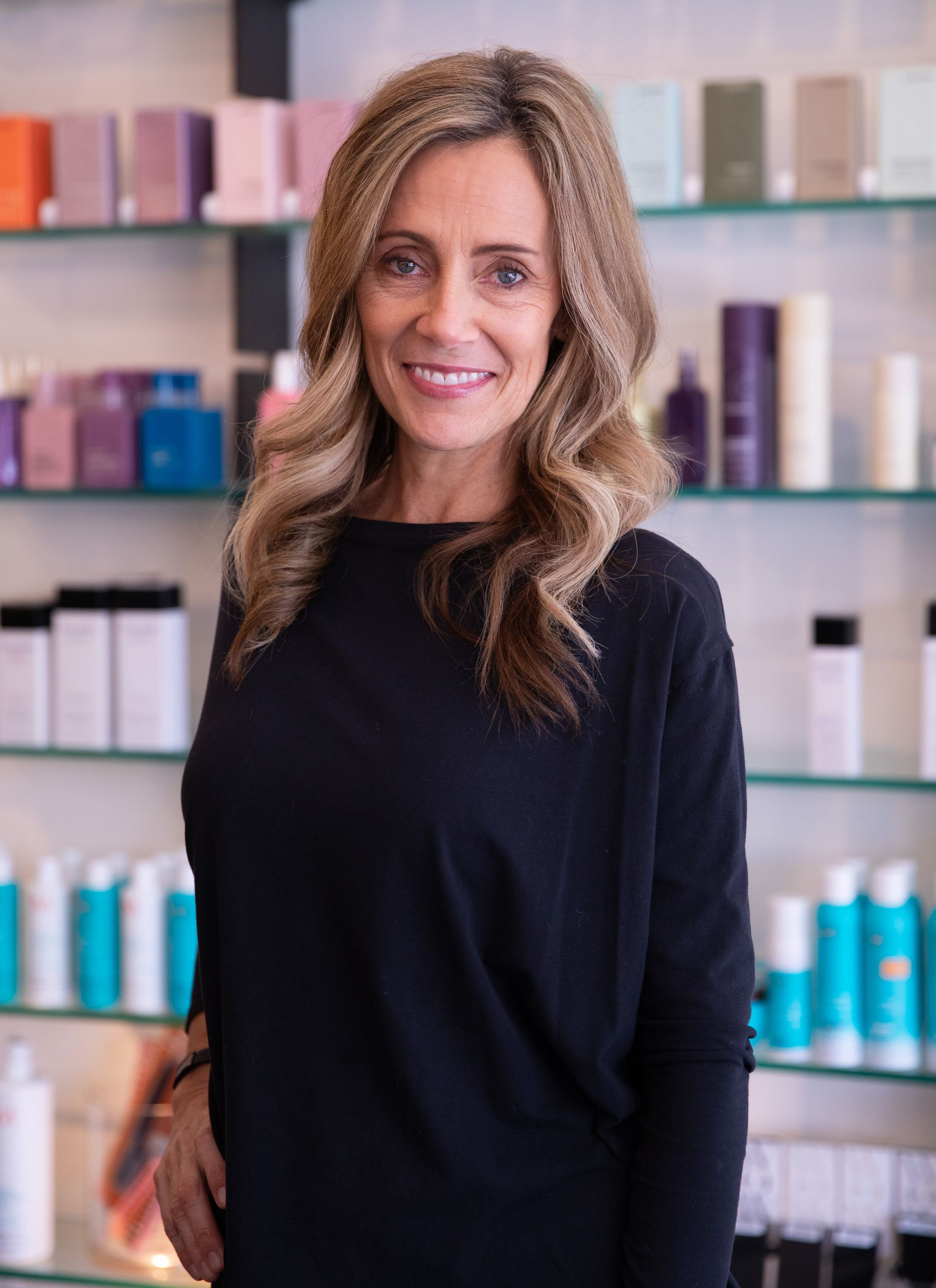 Woman in black sweater smiling in a salon, standing before shelves of colorful hair products