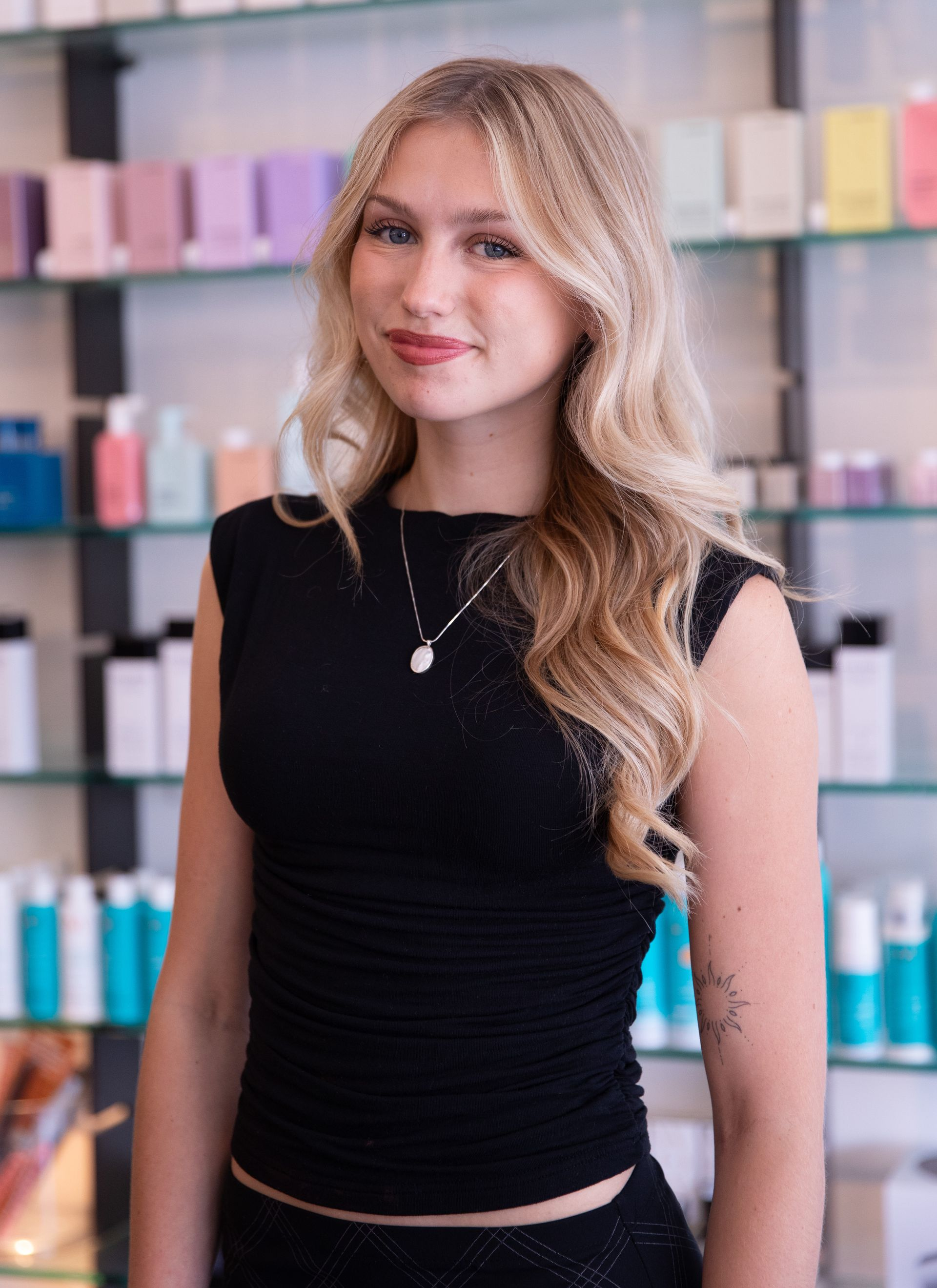 Woman in a black top standing indoors before shelves of colorful bottles, smiling at the camera