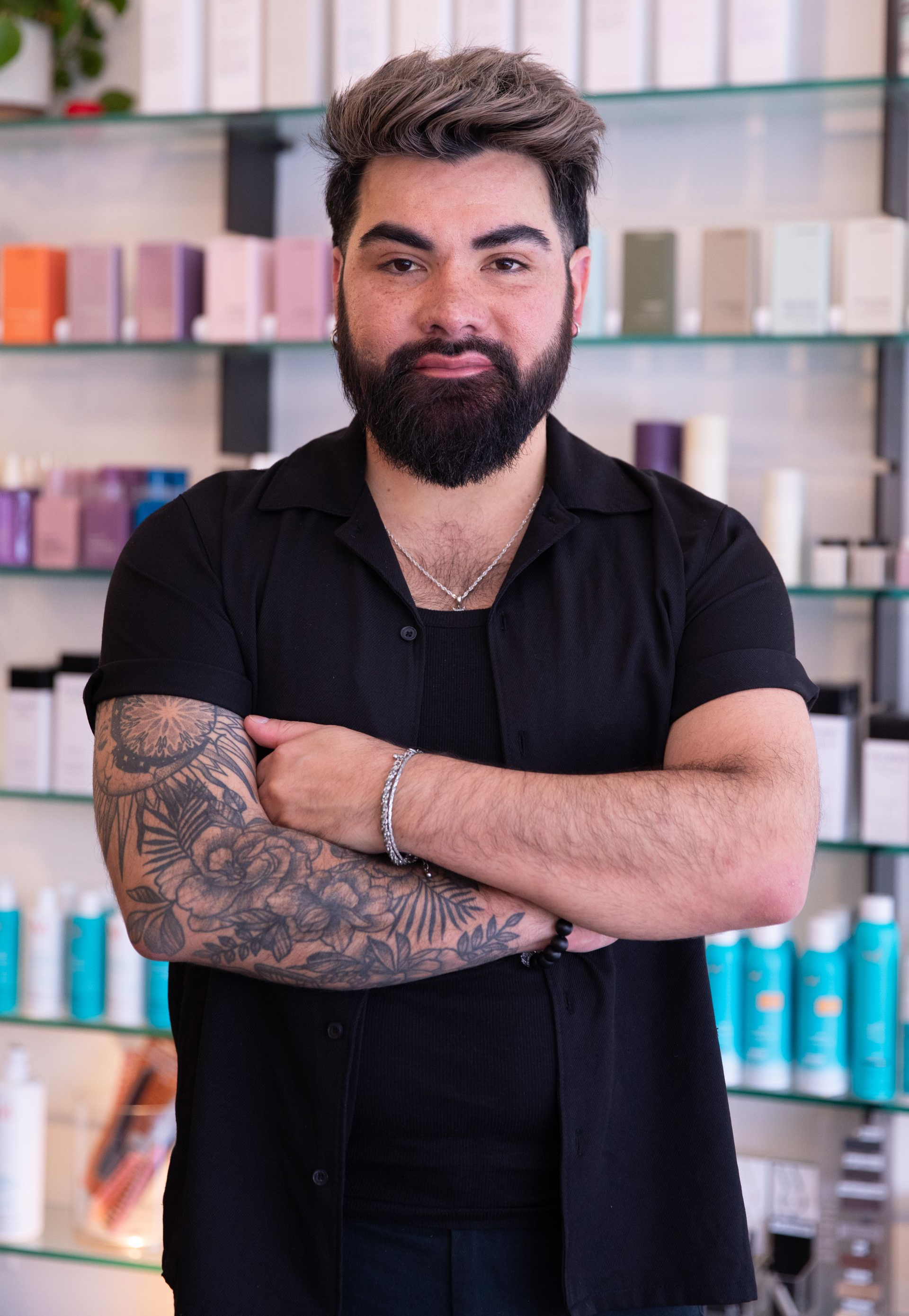 Bearded man with folded arms standing in a salon in front of shelves of hair products