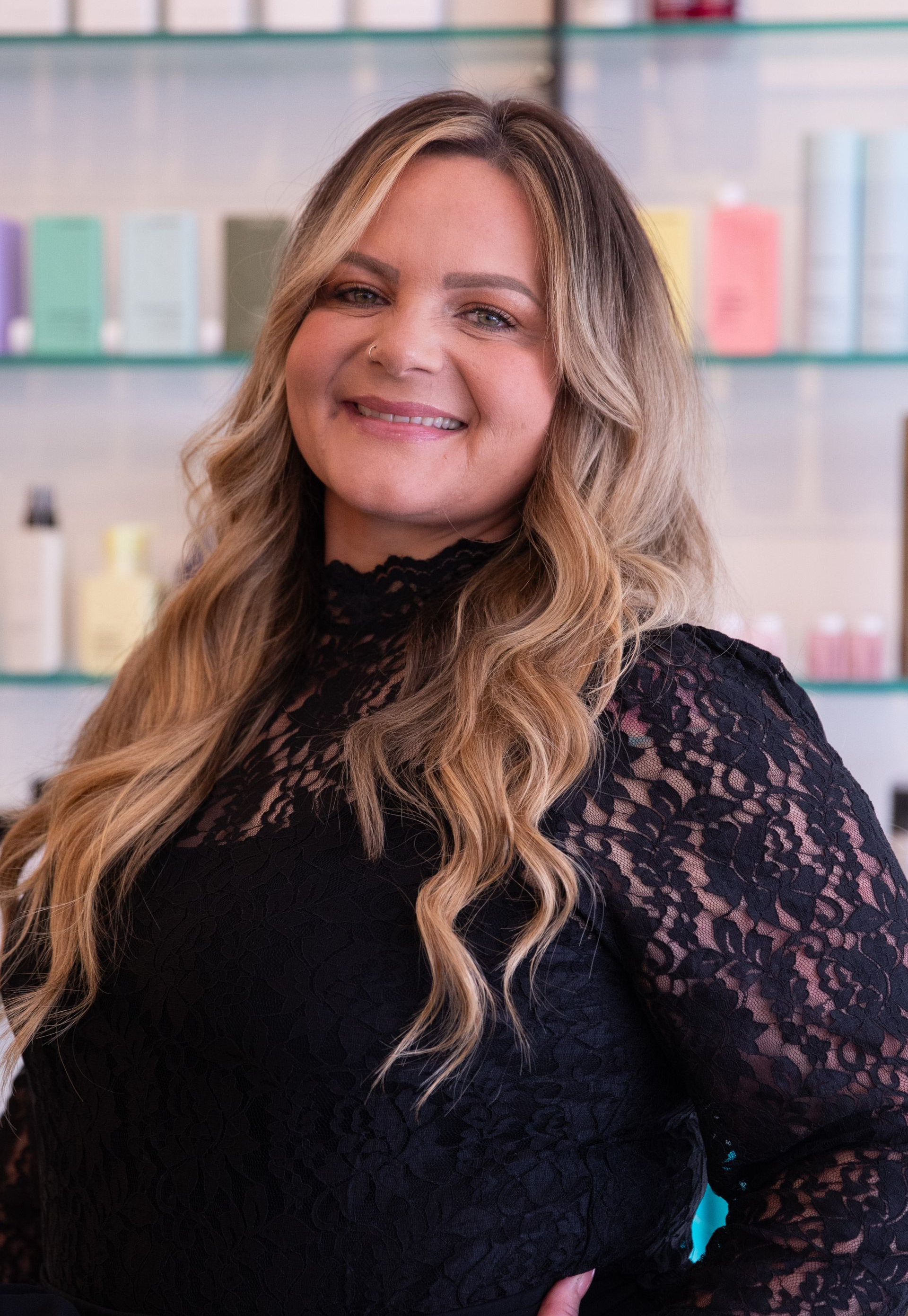 Smiling woman in a black lace top standing in front of pastel shelves in a salon setting