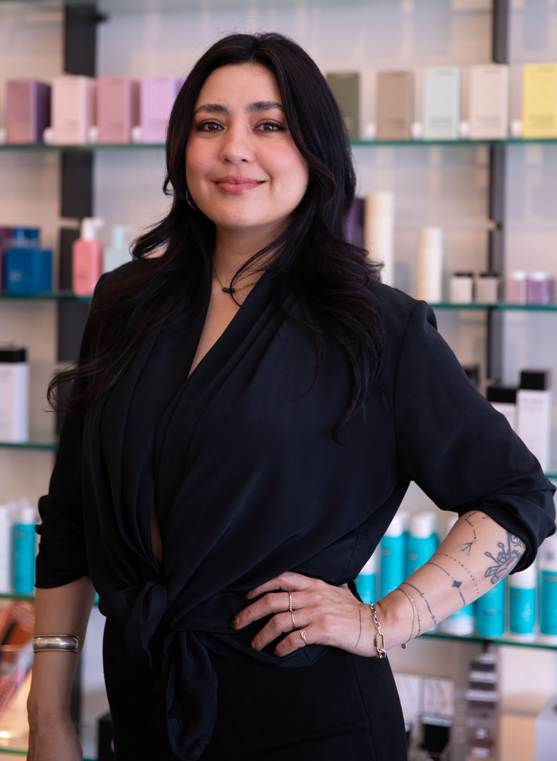 Woman standing in a salon with shelves of hair products, wearing a black outfit and smiling confidently