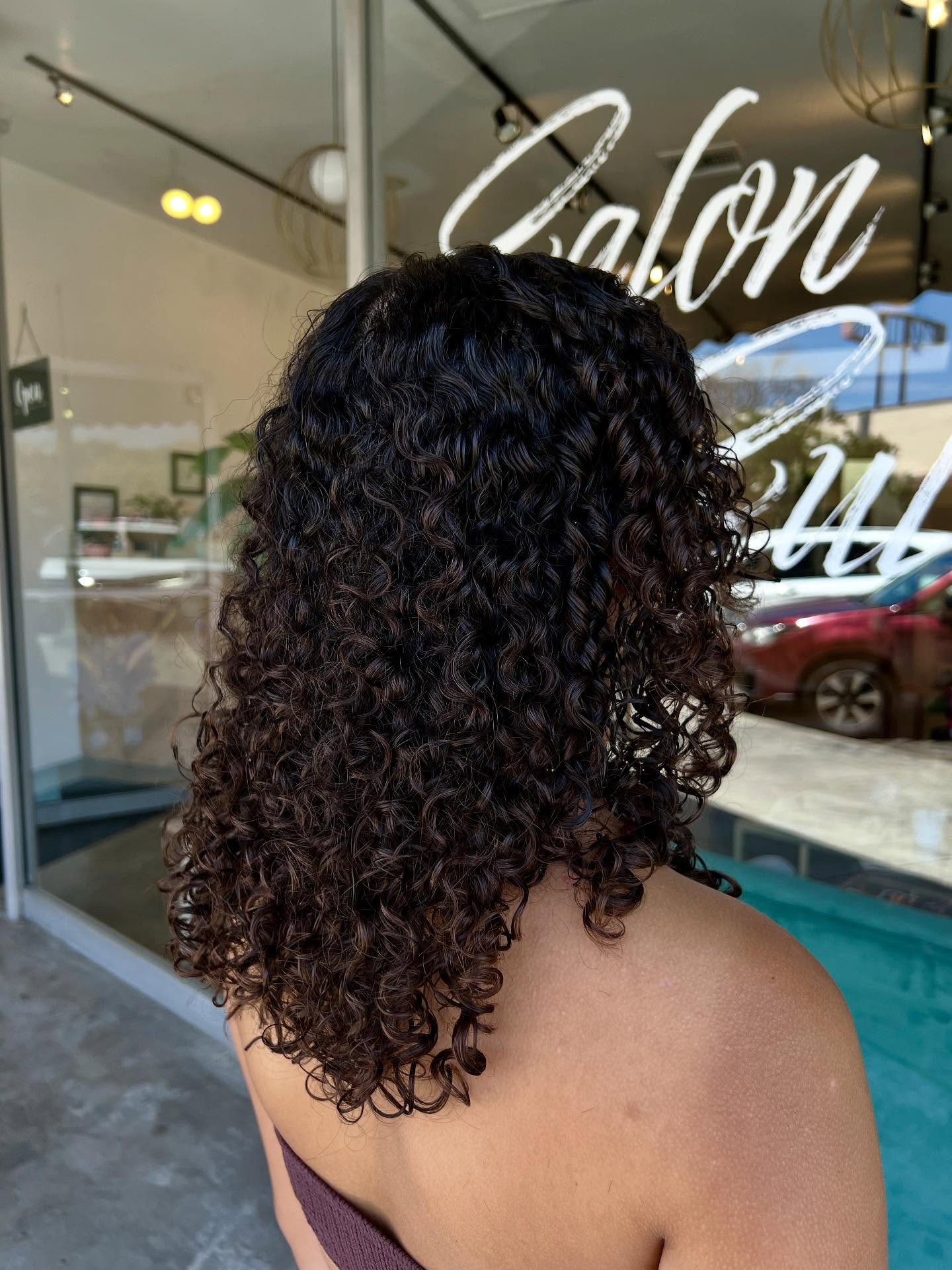Back view of curly dark hair at a salon storefront with glass windows and poolside reflections