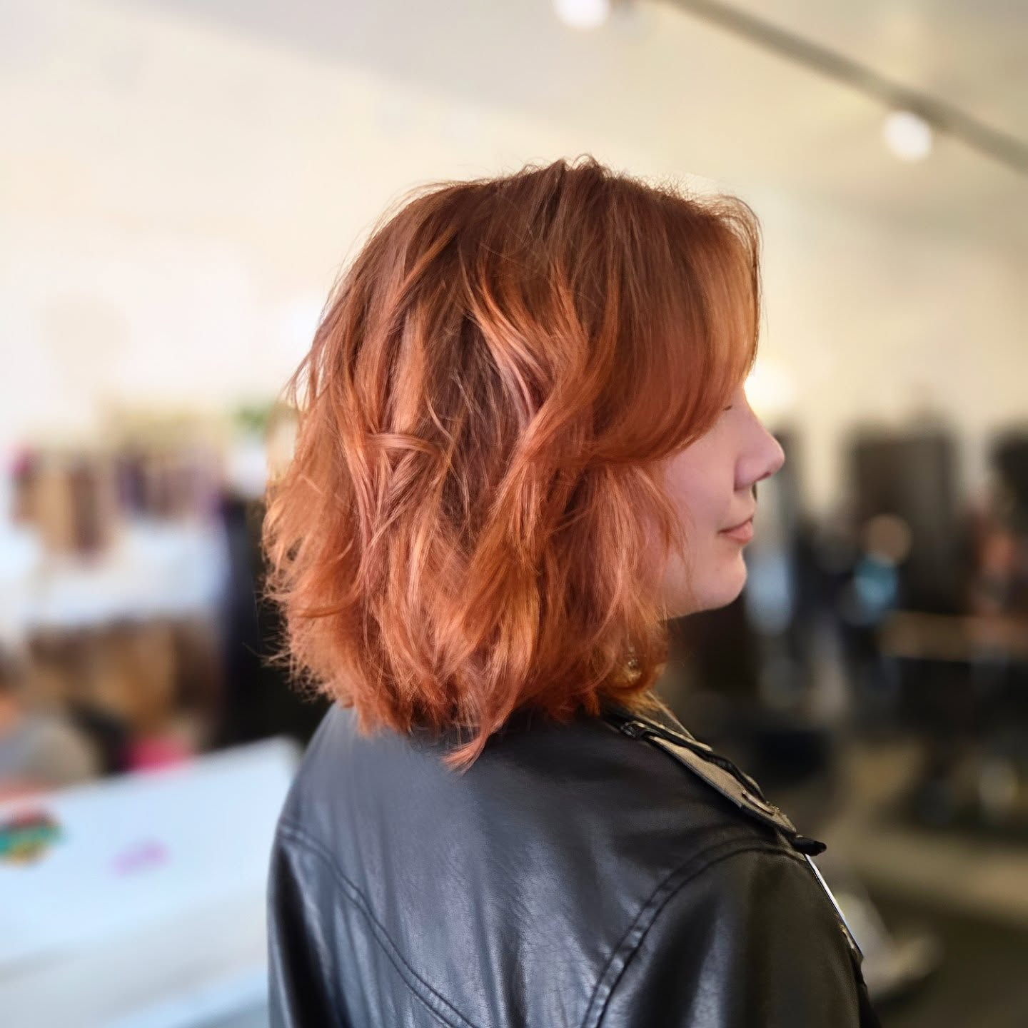 Side view of a person with wavy copper hair in a black leather jacket, indoors with a blurred background