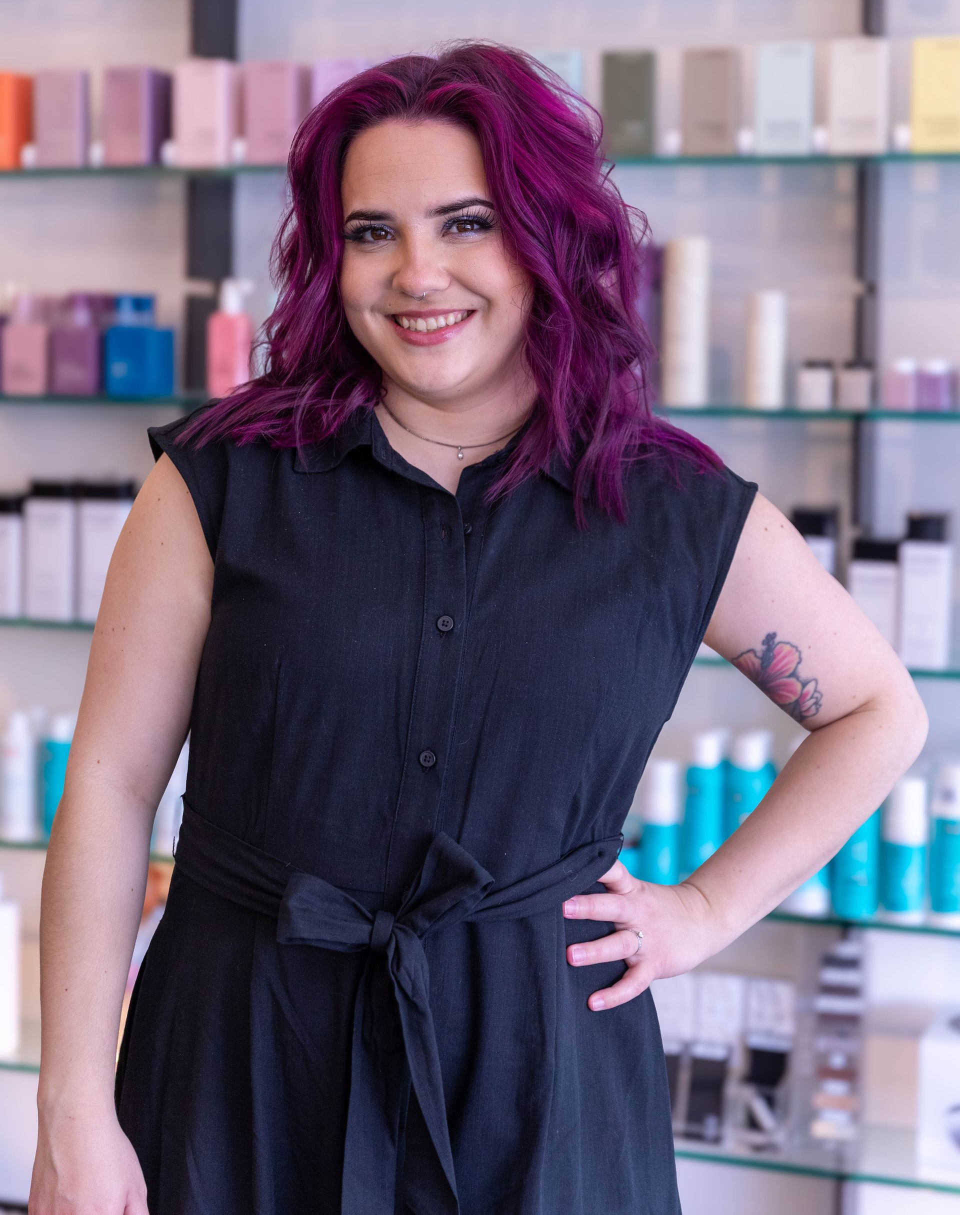 Smiling person with magenta hair in a black sleeveless dress, standing in front of colorful product shelves.
