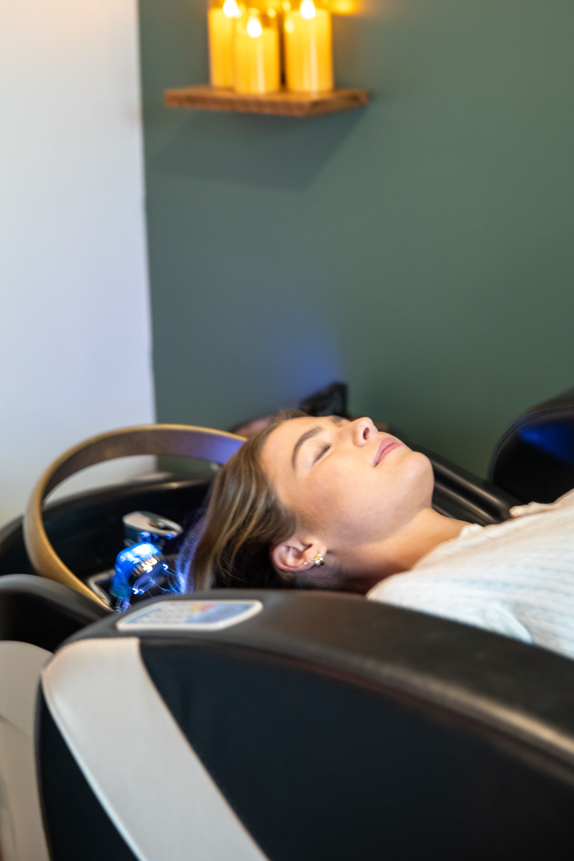 Person reclining at a shampoo sink in a salon, eyes closed, with lit candles on the wall nearby