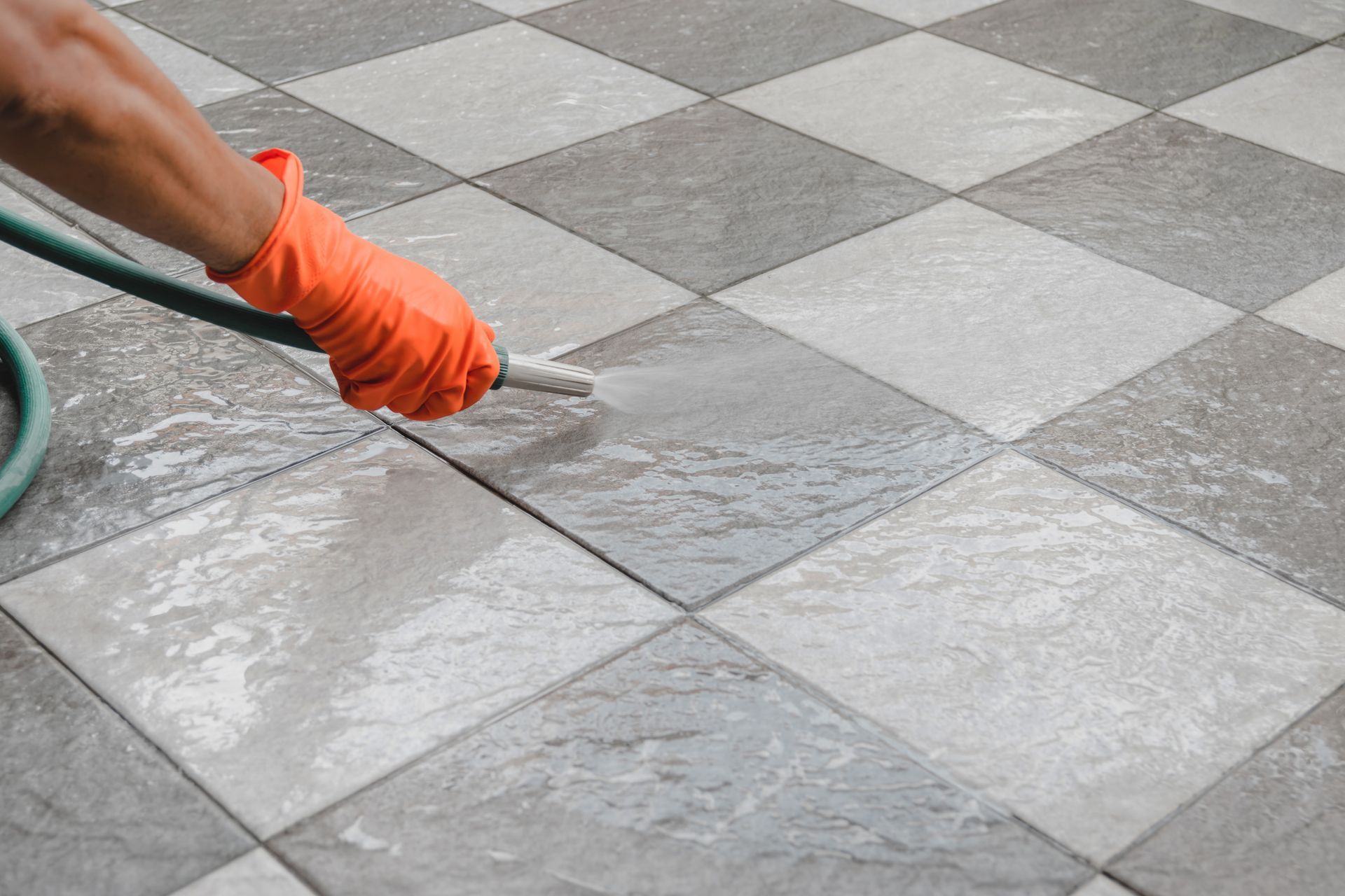 Person in orange glove spraying a checkerboard tile floor with a hose.
