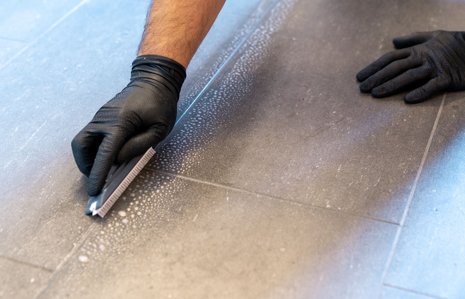 Person wearing black gloves cleaning floor grout with a tool.
