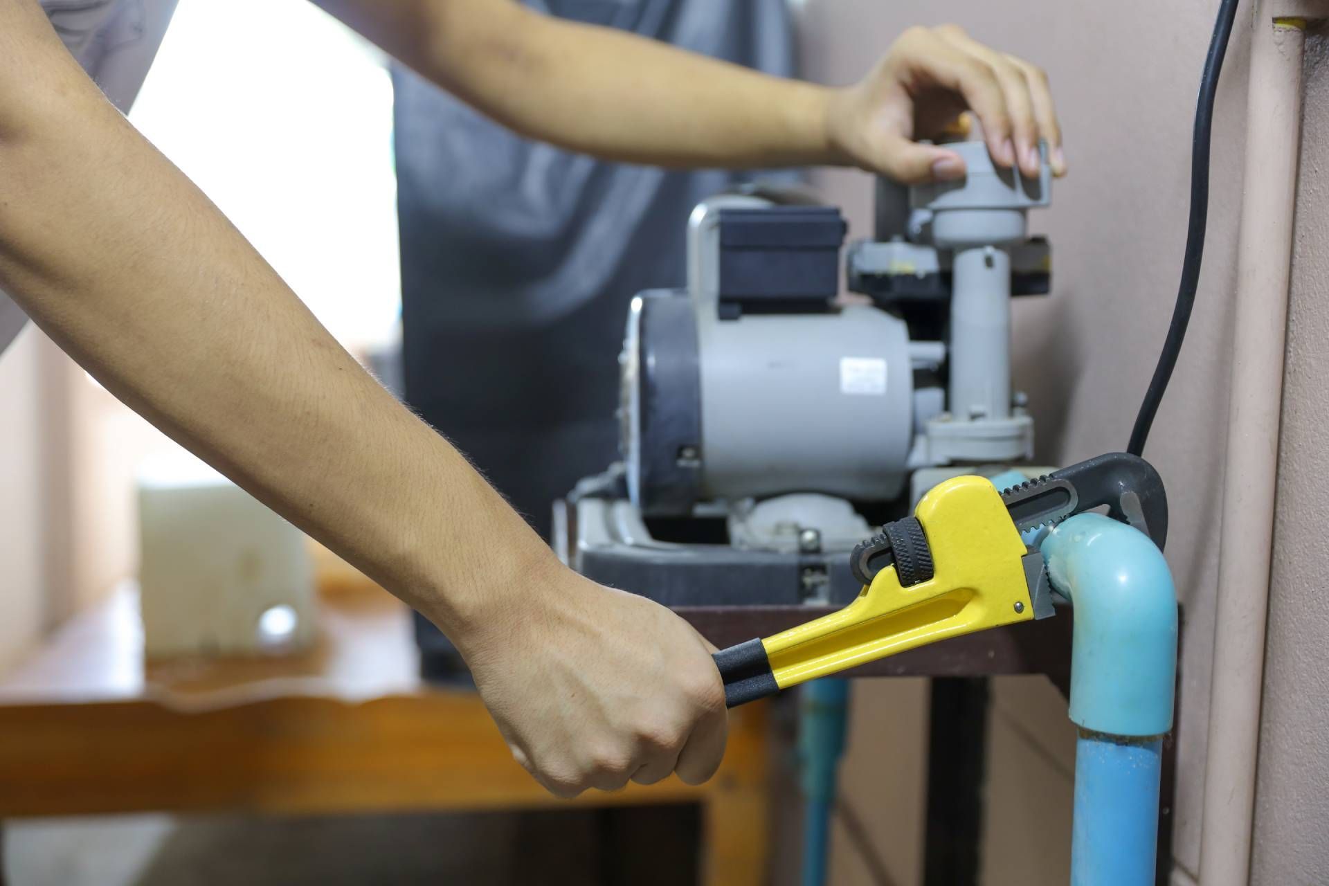 A person uses a yellow pipe wrench to tighten a connection on a blue PVC pipe attached to a grey water pump.