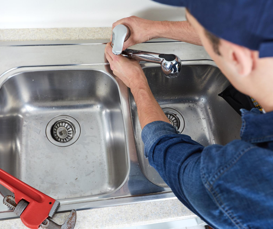 A plumber in a blue uniform repairing a kitchen faucet over a stainless steel double sink.
