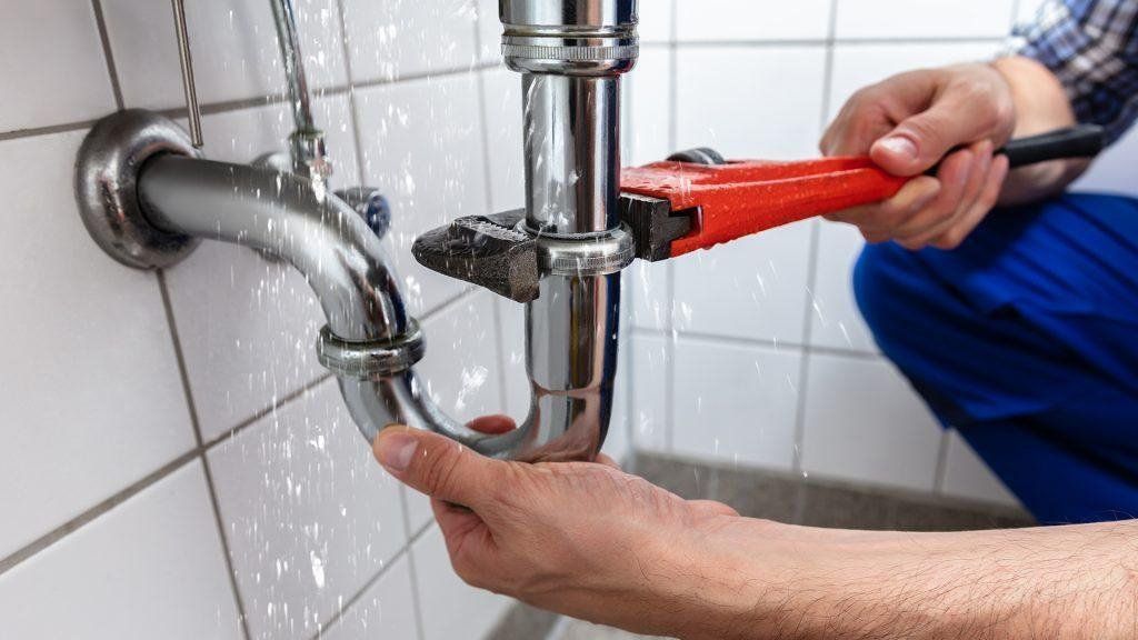 A plumber in blue work pants uses a red pipe wrench to tighten a leaking chrome P-trap under a sink with white tiled walls.