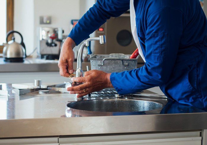 A person in a blue uniform repairing a stainless steel kitchen faucet over a sink.