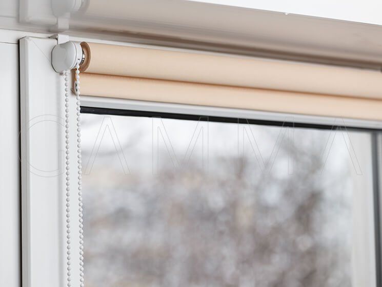 Tenda a rullo beige che copre parzialmente una finestra, cornice bianca, catena, sfondo esterno sfocato.