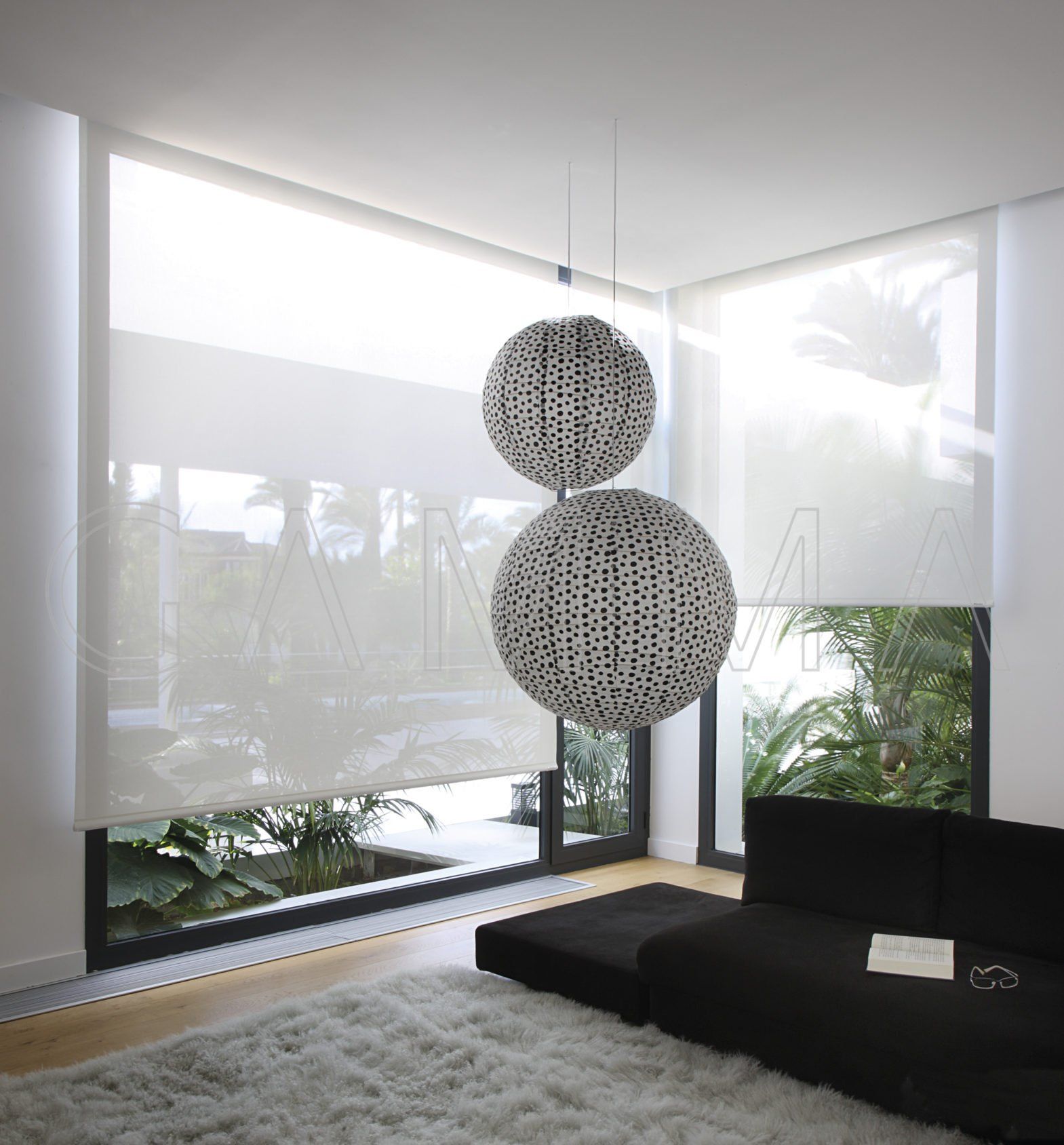 Stanza con ampie finestre, persiane bianche, divano nero e sfere decorative sospese.