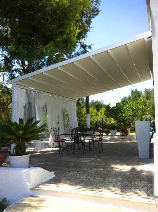 Patio esterno con tenda da sole retrattile sopra un tavolo da pranzo, tende trasparenti e piante in vaso.