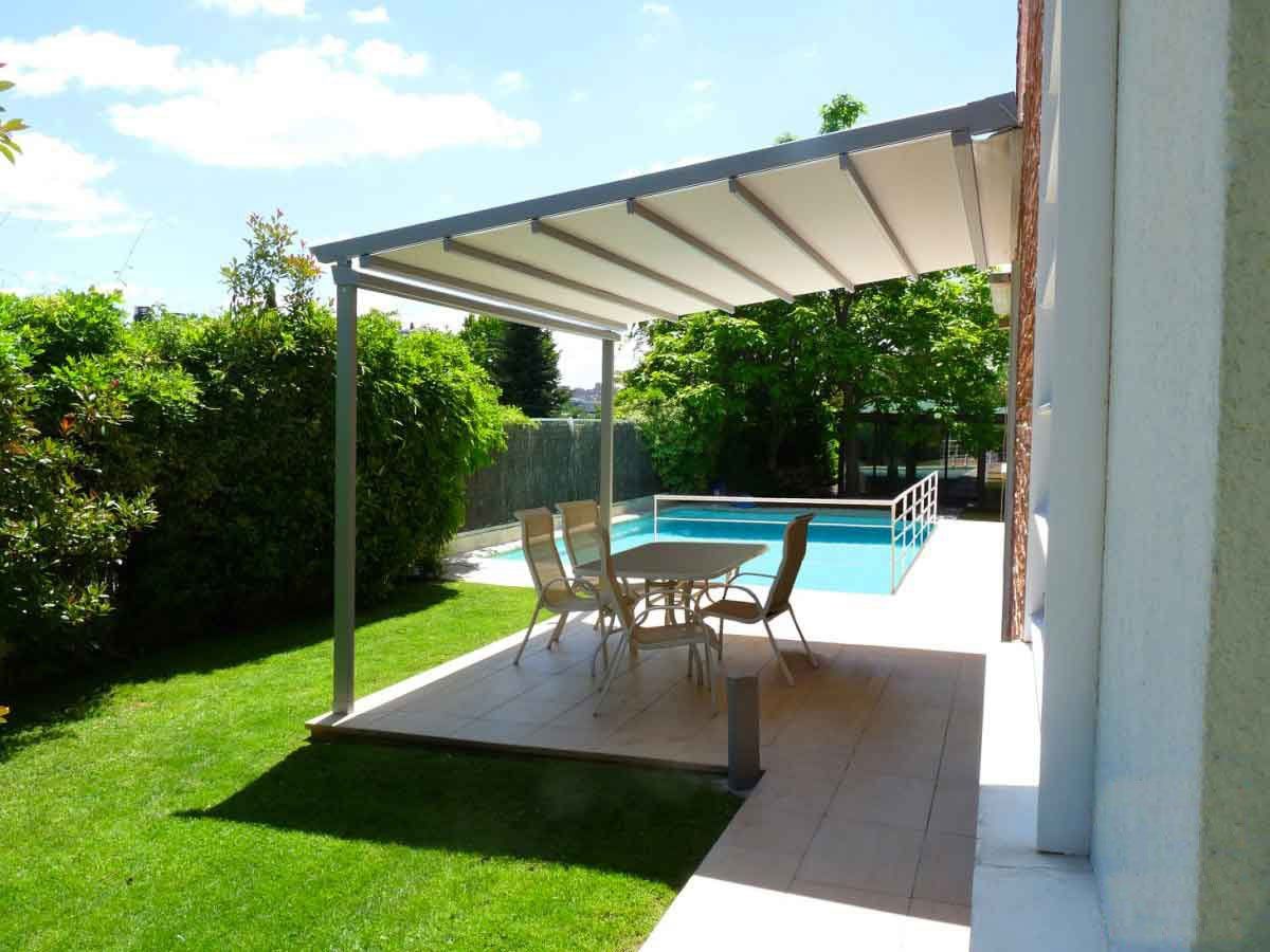 Patio con pergolato, tavolo, sedie e piscina. Erba verde, acqua azzurra e tenda bianca. Giornata di sole.