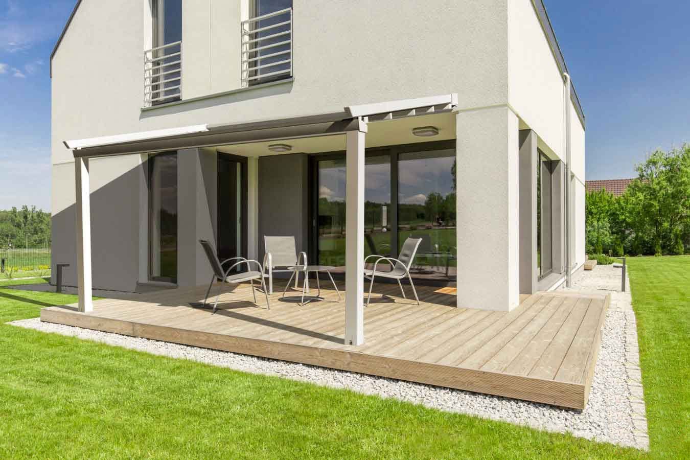 Casa moderna con terrazza in legno, mobili da giardino e pergolato, circondata da prato verde.