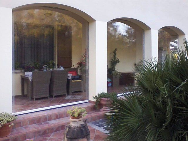 Patio esterno con archi, racchiuso da teli trasparenti. Tavolo e sedie visibili all'interno.