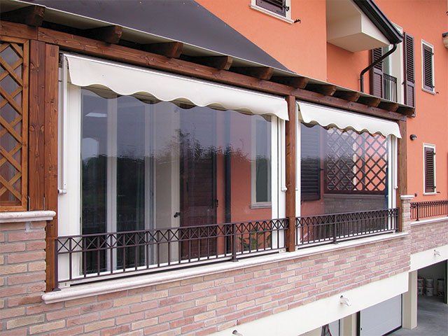 Balcone con finestre in vetro e tende da sole, struttura in legno marrone, muro in mattoni, ringhiera in ferro battuto.