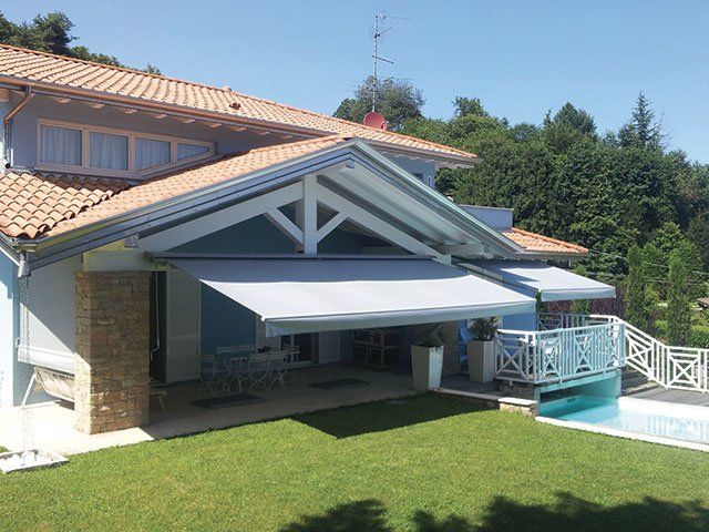 Casa blu con tende da sole grigie, tetto di tegole e piscina in una giornata di sole.
